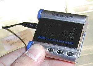 コンパクトな携帯プレーヤー『SV-SD70』。幅48×奥行き15×高さ46mmというサイズ。中央部分が12×2行の文字表示部。その上にも1行分の情報表示部がある。単4電池は上の筒状の部分に格納する 